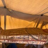 A golden beige Egyptian coffin on two wooden risers with the feet of the left side and the head on the right side. The coffin is decorated with a variety of green, black and red images and inscriptions. The coffin is sitting in a low-hanging canvas tent.