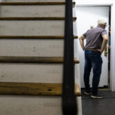 Man wearing hat in an apartment building hallway speaks to man behind a door narrowly opened.