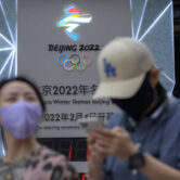 People wearing face masks walk past a display showing a countdown clock to the 2022 Winter Olympics in Beijing.