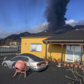 Ash covers a house, car and garden as in the background a volcano erupts on the Canary Island of La Palma, Spain.