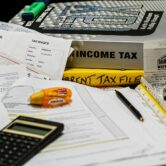 A calculator and stationery supplies sit on a desk with books about income tax
