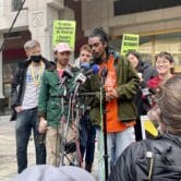 a small group of Amazon employees stand outside the NLRB office in downtown Brooklyn in front of microphones. They are surrounded by journalists holding microphones and taking notes
