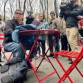 Amazon Labor Union organizer Jason Anthony sits in a bright red chair at a bright red table looking disappointed. A group of reporters with cameras and notepads stands behind him.