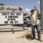 A person takes a photo in front of a sign referencing the extreme heat in Texas.