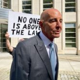 Tom Barrack wearing a grey suit standing outside of the federal courthouse in Brooklyn, behind him a man is standing holding a sign that says in all capital letters "no one is above the law"