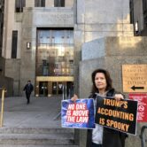 Woman holds signs saying "NO ONE IS ABOVE THE LAW" and "TRUMP'S ACCOUNTING IS SPOOKY" in front of the gray stone Manhattan criminal courts building
