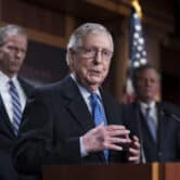 Mitch McConnell, standing in front of two other men, gestures behind a lectern while speaking to reporters.