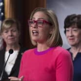 Kyrsten Sinema stands in front of Tammy Baldwin and Susan Collins during a press conference.