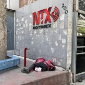 A red Notimex logo hangs on a dilapidated grey wall. Below it a homeless man sleeps wrapped in a red blanket