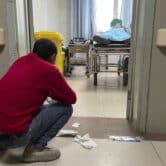 A man looks inside a treatment room where an elderly person receives help with breathing via a manual ventilator pump.