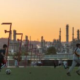 Children playing soccer with an oil refinery in the background.