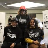 Two people who are seated hold signs that read "CA Reparations Now 2023," while another person stands behind and between them.