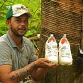 A rubber tapper holds up a pair of Veja sneakers in front of a tree in the Amazon rainforest.