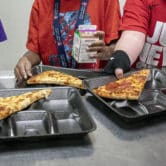 Three students select their meals, which include slices of pizza and chocolate milk, during a lunch break in an elementary school cafeteria.