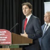 Justin Trudeau gestures while speaking behind a lectern, with Doug Ford standing in the background.