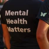 Two women stand on either side of a woman wearing a shirt that reads "Mental Health Matters" on the back.