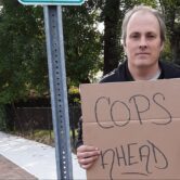 a man stands on the sidewalk holding a cardboard sign that says "cops ahead" in all capital letters written in black marker