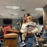 A woman stands in front of a podium and a microphone holding her young grandson in her arms