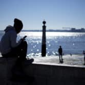 A young man sits while checking his phone by the Tagus river.