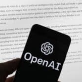The OpenAI logo is seen on a mobile phone in front of a computer screen displaying text.