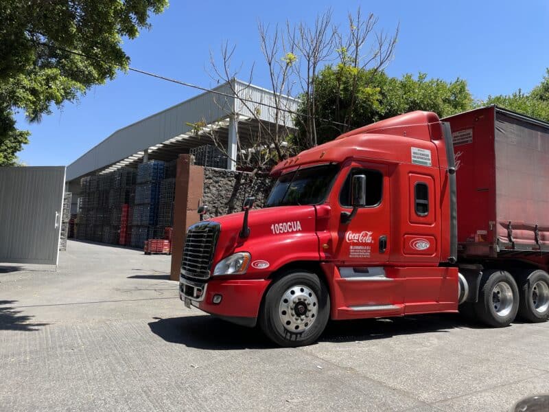 A bright red semi truck is parked in front of the entrance to a warehouse. Bottles of water and soda pop are stacked up in the warehouse