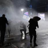 Israeli police use a water cannon to disperse demonstrators blocking a road. A protester kneels while holding up the Israeli flag.