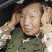 Iwao Hakamada holds his hands over his ears while sitting in a car.
