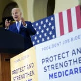 Joe Biden gestures while delivering a speech on a stage with Medicare-related signs around him.