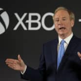 Brad Smith gestures while speaking on stage in front of a sign with the Xbox and Microsoft logos.