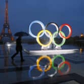 A man walks past the Olympic rings as it rains in Paris. The Eiffel Tower is in the background.