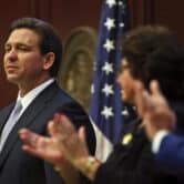 Two people applaud while Ron DeSantis stands next to a microphone, with the American flag in the background.