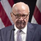 Rupert Murdoch stands in front of two American flags during an event.