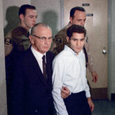 Russell E. Parsons holds Sirhan Sirhan's arm as they and two guards walk in a courthouse hallway.