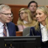 Steve Owens and Gwyneth Paltrow smile while sitting in a courtroom, with several people seated behind them.