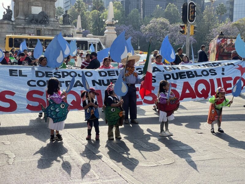 Five small children wearing colorful cardboard fish outfits stand in front of a banner with Spanish text on it. People behind the banner hold baby blue water droplets