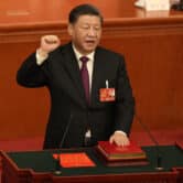 With his left hand on a book, Xi Jinping raises his right fist while standing behind a lectern at China's Great Hall of the People.