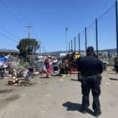 A police officer, foreground, watching a group of people and some belongings on the ground.