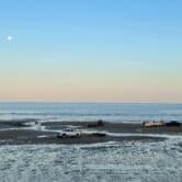 Three pickup trucks drive on a beach exposed by low tide. One hauls a boat. The blue sky with the moon in the upper left corner dominates the upper half of the foto