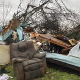 Snapped trees and damaged household items, including a recliner, on a grass field.