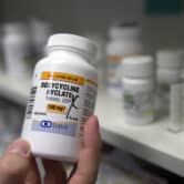 A personal holds a bottle of doxycycline hyclate at a pharmacy, with bottles of pills on shelves in the background.