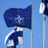 A NATO flag and two Finnish flags blow in the wind.