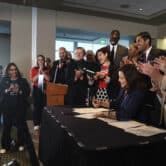 Gretchen Whitmer signs a package of gun bills as people applaud while standing around her.