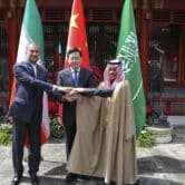 Hossein Amirabdollahian, Prince Faisal bin Farhan Al Saud and Qin Gang shake hands in front of Iranian, Chinese and Saudi Arabian flags.