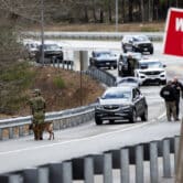 Law enforcement officials, including one holding an assault rifle as another one guides a dog on a leash, investigate a shooting on a highway.