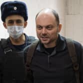 A man wearing a face mask escorts Vladimir Kara-Murza, who is wearing a backpack.