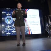 William Hartman gestures while speaking on stage, with a screen showing the U.S. Cyber Command insignia in the background.