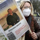 A woman wearing a white KN95 face mask holds a poster featuring a photo of Benjamin Briere.
