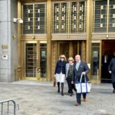 E. Jean Carroll and Roberta Kaplan are walking out of the revolving doors at Manhattan federal court building, a man next to them is holding a tote bag