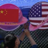 A man attaches signs to a canopy. One sign reads "China" with the Chinese flag, while the other reads "America" and features the United States flag.