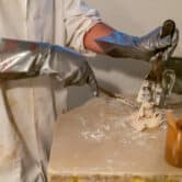 A scientist in metallic silver gloves using two spatulas to mix a beige, clay-like substance on a tan table.